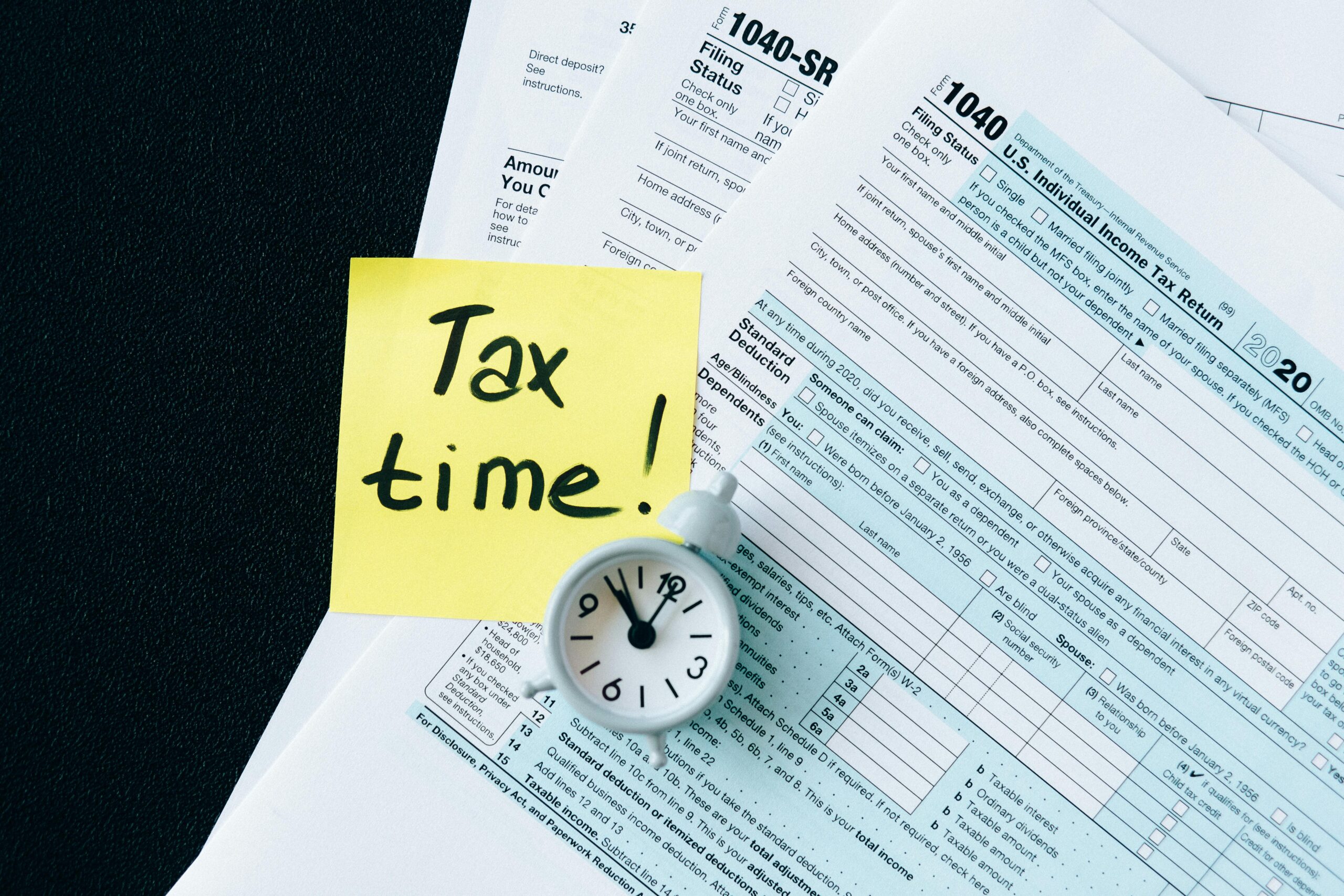 A stack of tax forms with a clock and yellow sticky note saying 'Tax time!' indicating urgency.