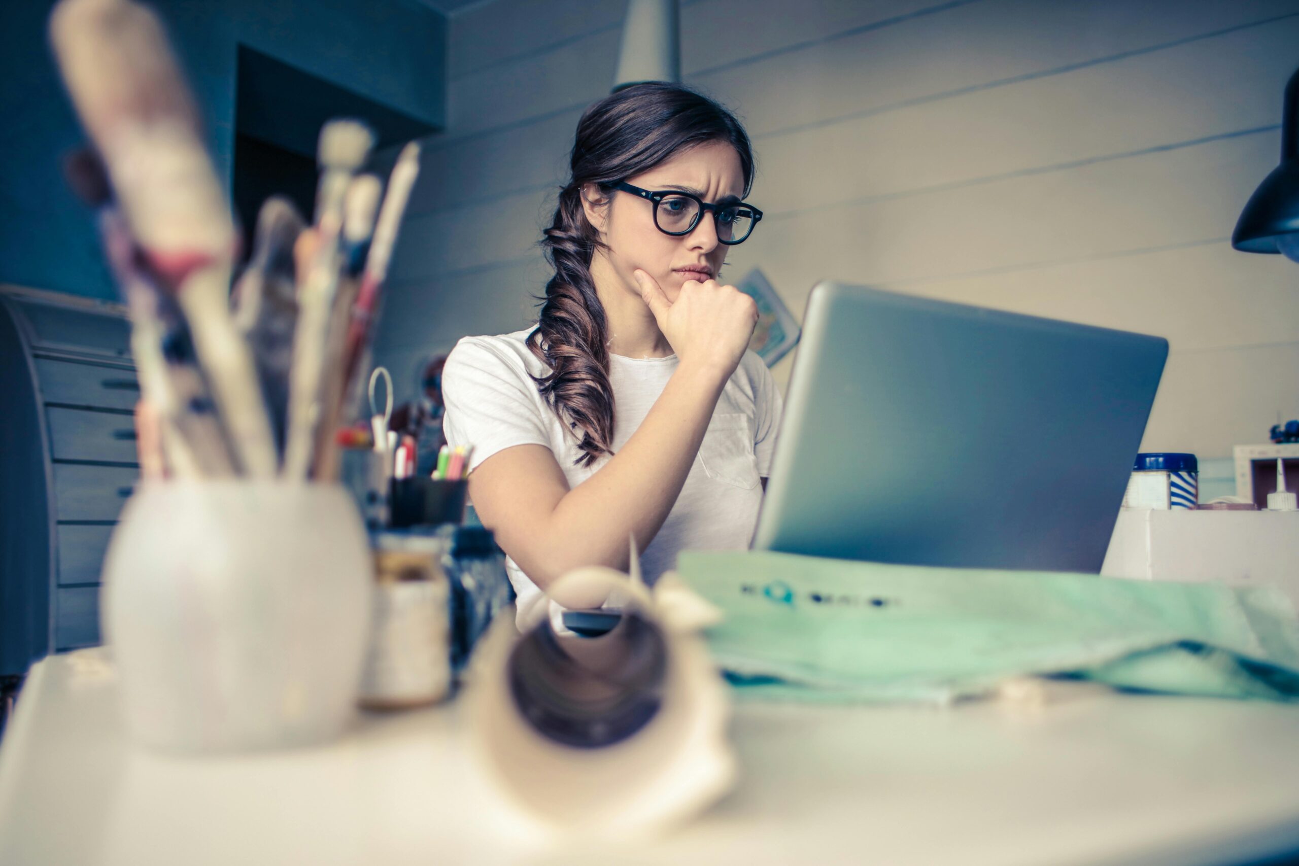 Young woman with glasses deeply focused on a laptop surrounded by art supplies in a home office.